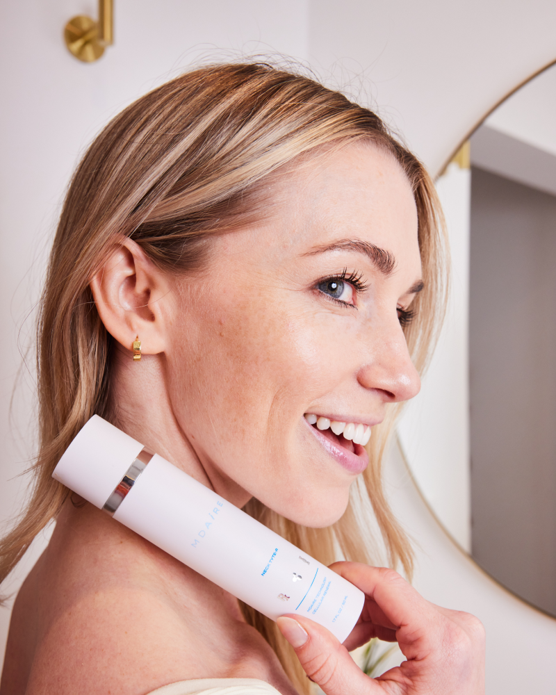 Woman holding the MDAiRE Neck Tyte-R skincare product bottle, posing in a bathroom setting