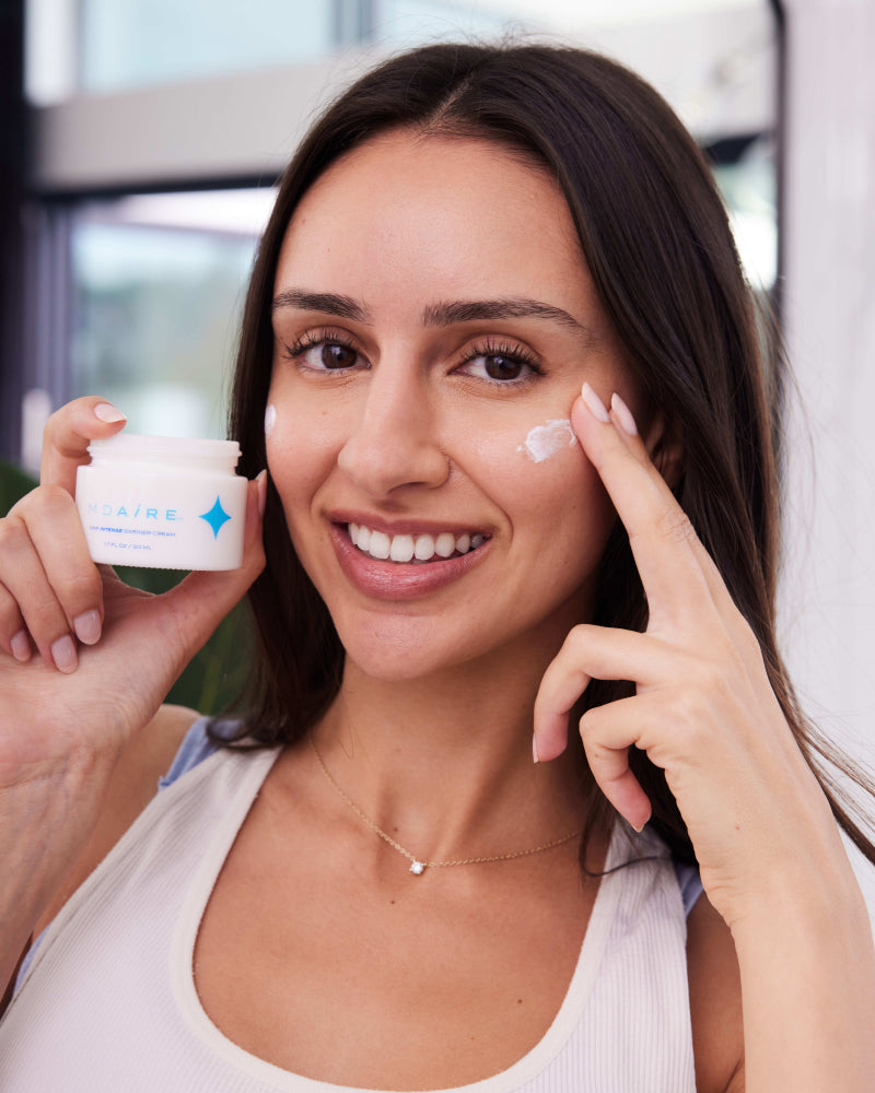 Woman with brunette hair applying MDAiRE NMF Intense Barrier cream to her face with a jar of the Barrier cream in her hand.