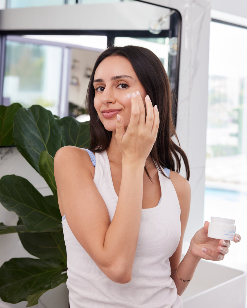 Dark-haired woman applying NMF Intense Barrier cream to her face in a bright room with plants