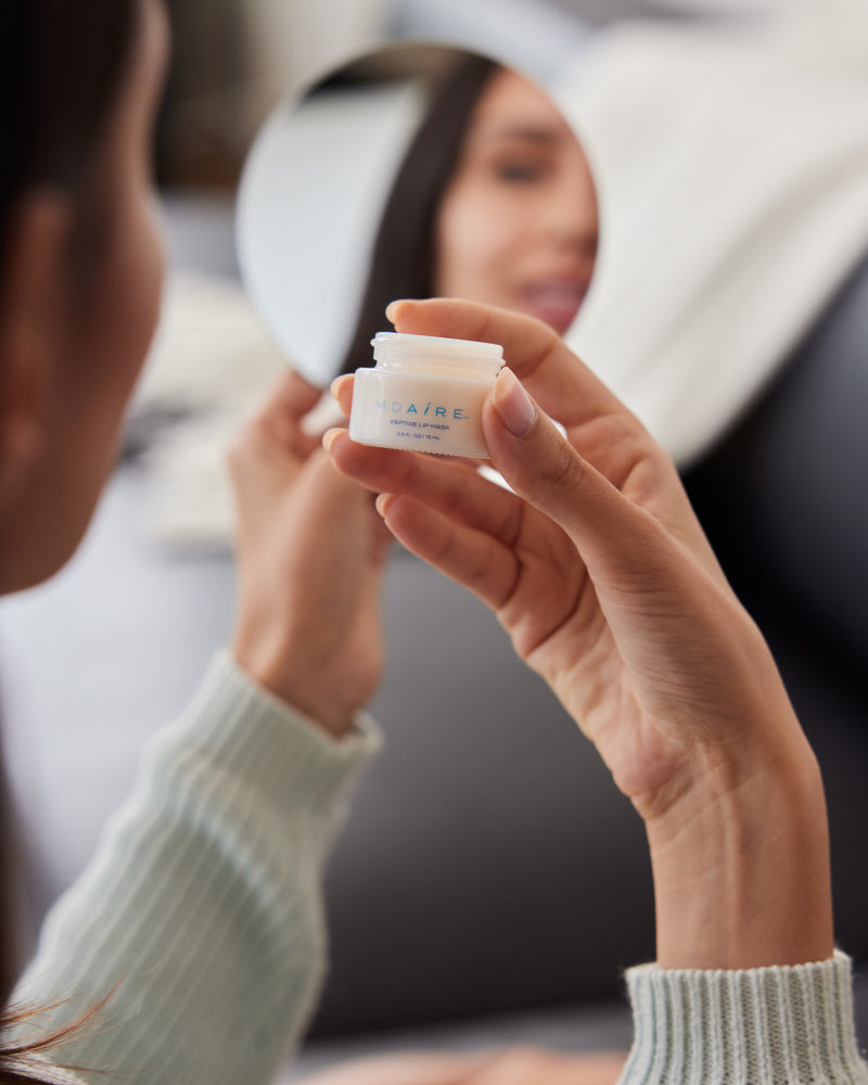 Woman looking into a mirror, holding a MDAiRE peptide lip mask skincare product with a blurred background